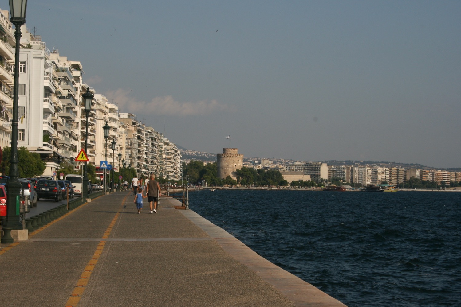 a sidewalk next to a body of water with Thessaloniki in the background