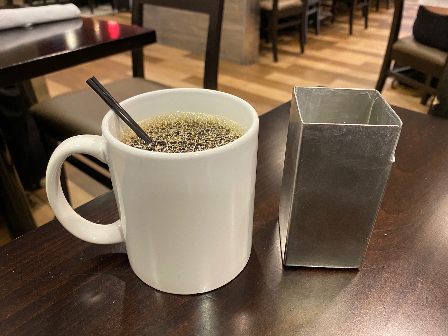 a white mug with brown liquid and a metal container on a table