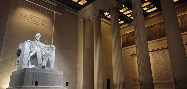 a statue of a man sitting on a chair in a large room with columns