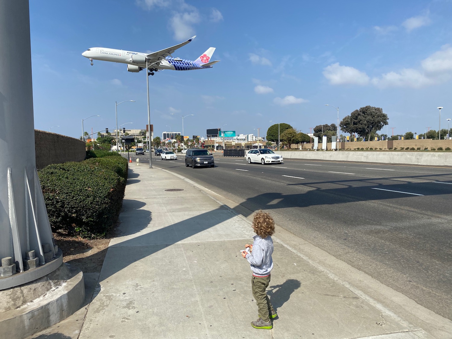 a child standing on a sidewalk looking at an airplane flying over a street