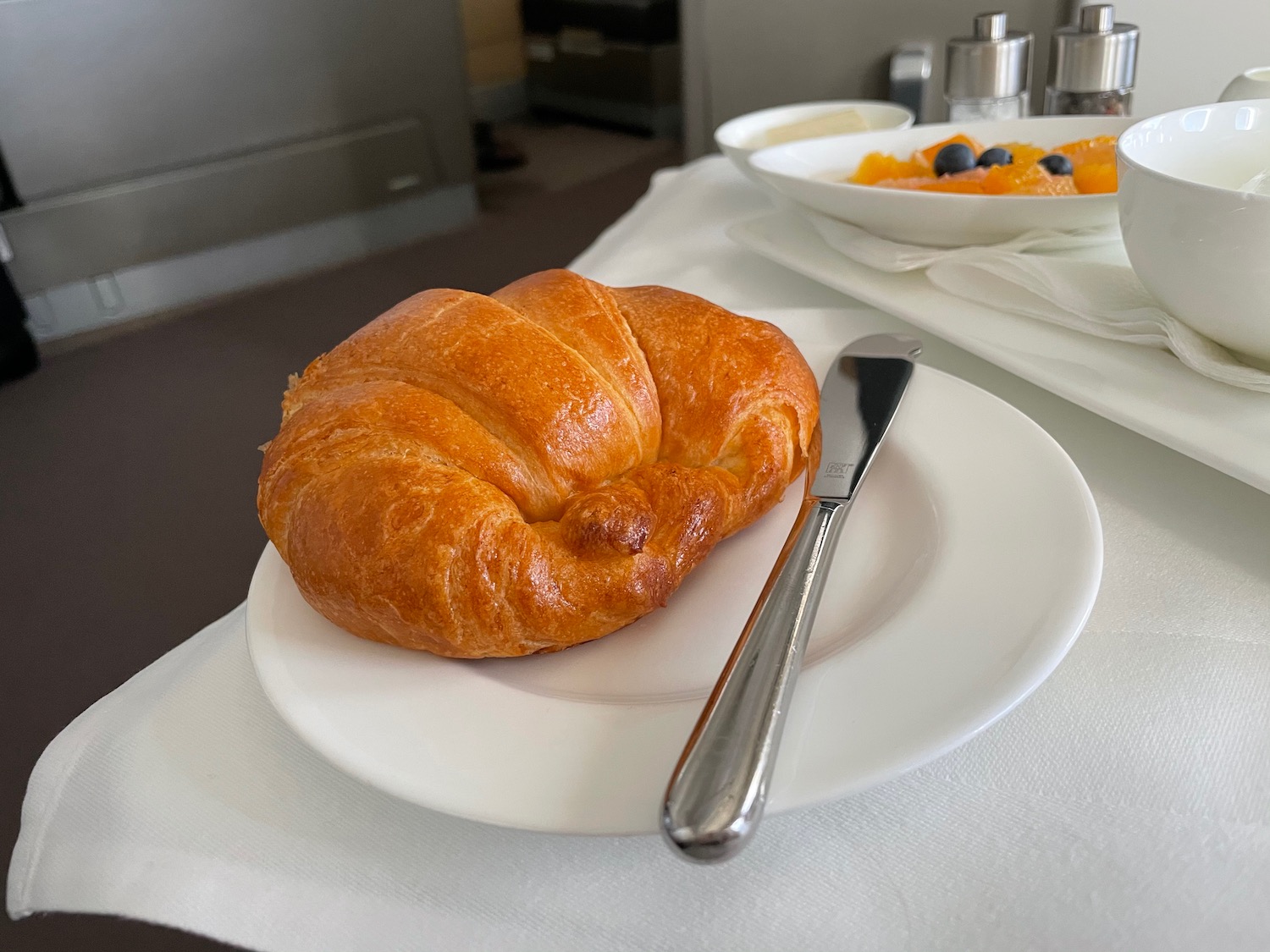 a croissant on a plate with a knife and a bowl of fruit