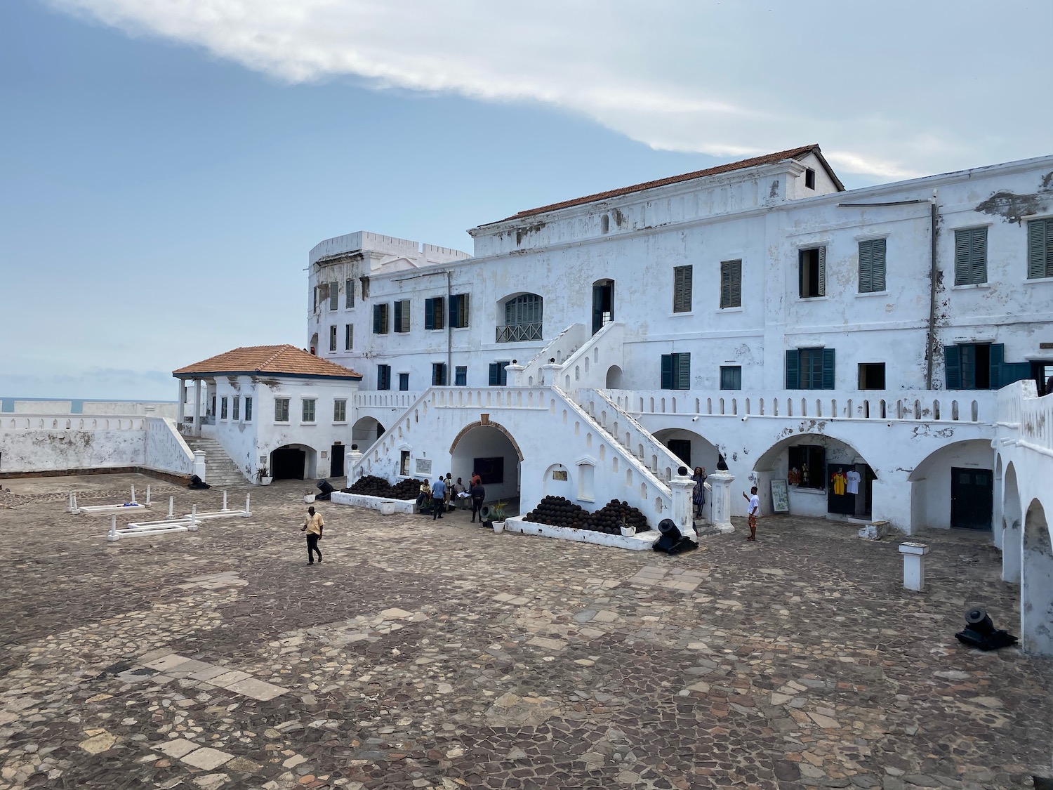 a white building with stairs and people walking around with Cape Coast Castle in the background
