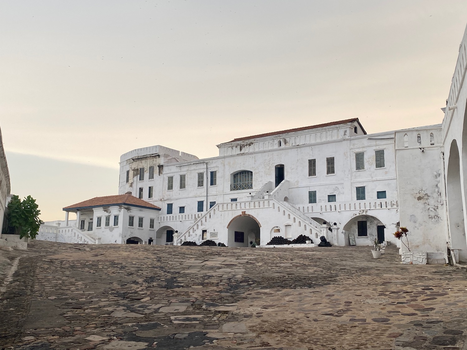 a white building with stairs and a stone walkway with Cape Coast Castle in the background