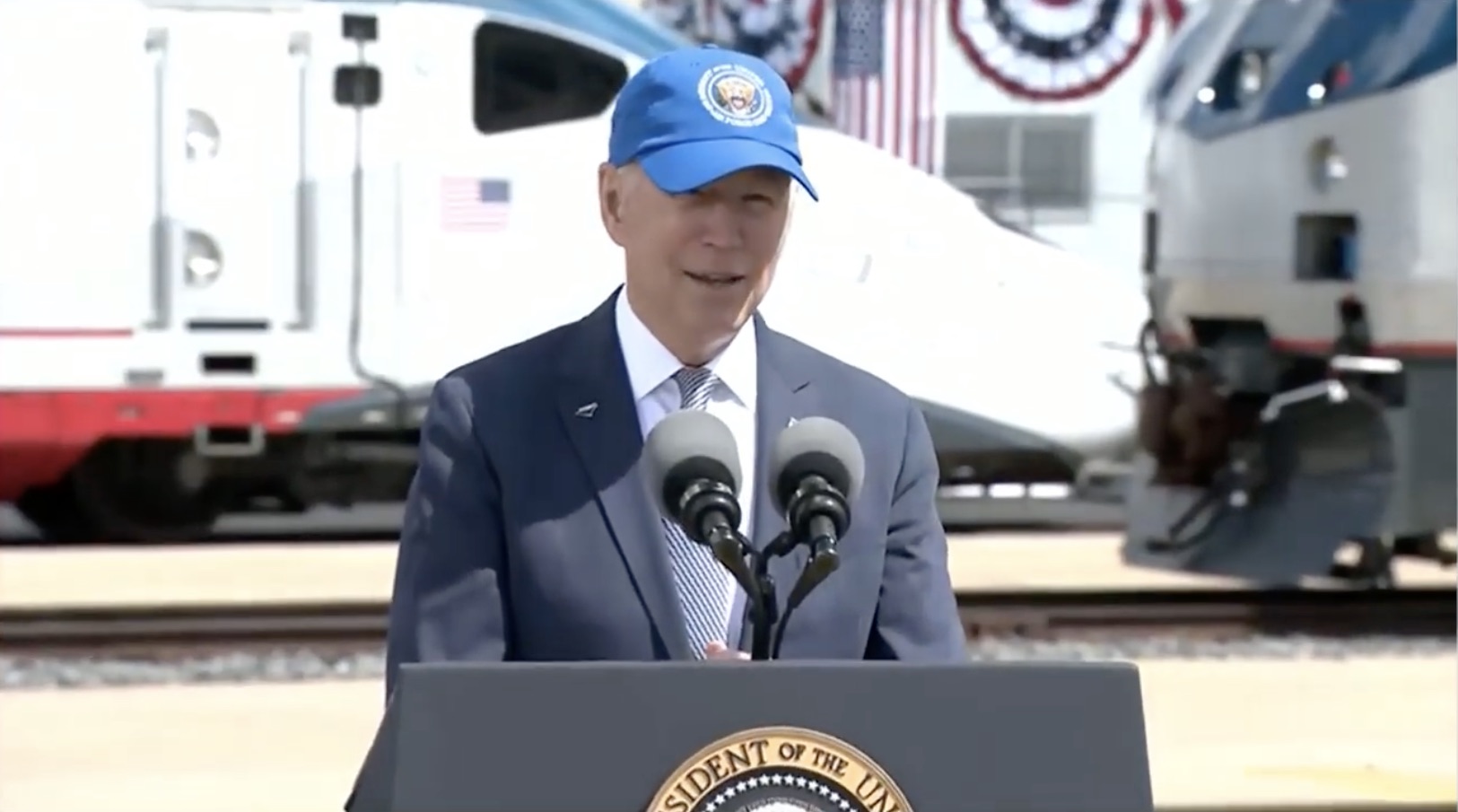 a man in a blue hat and suit standing at a podium with microphones