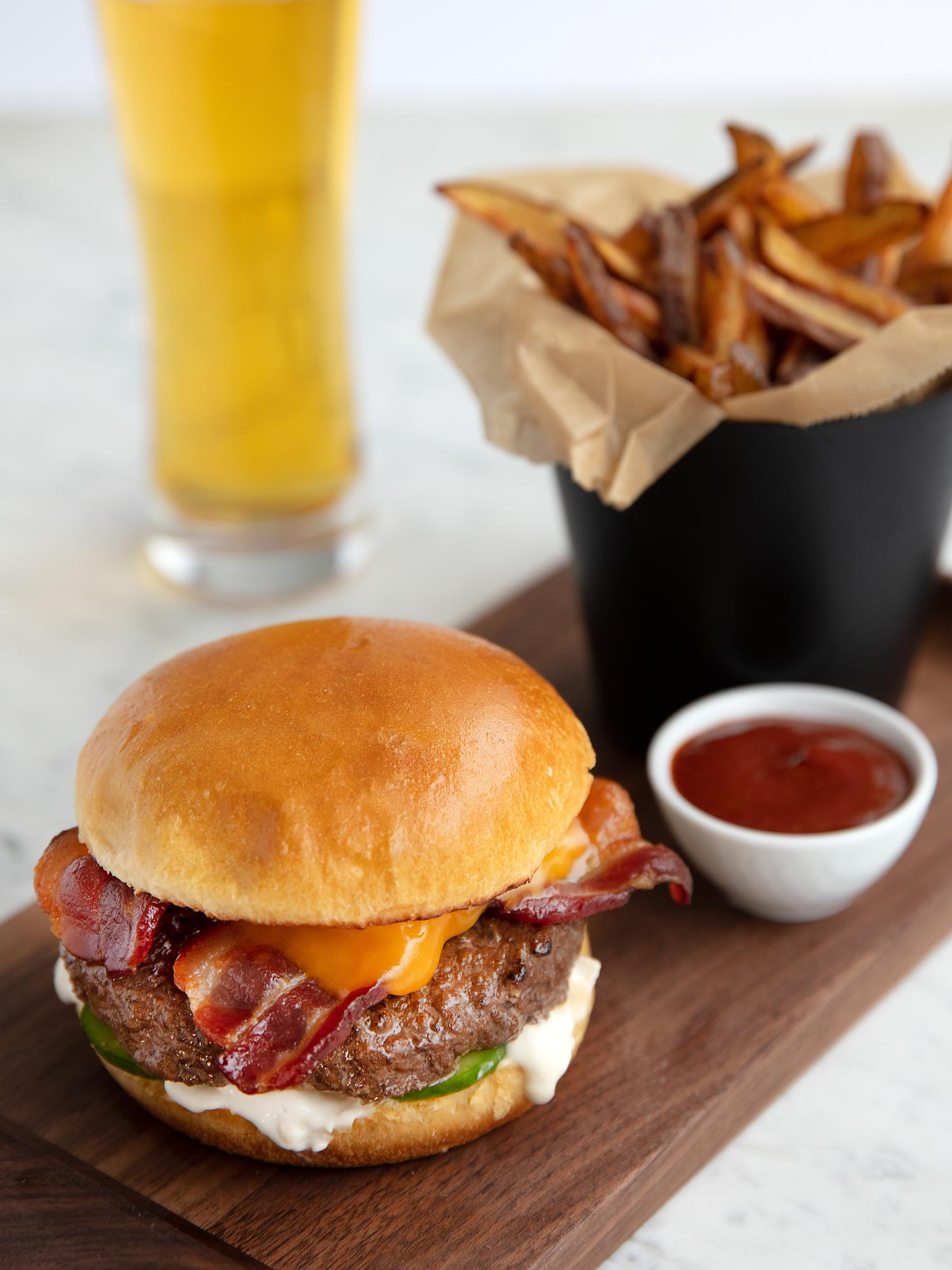 a burger with bacon and cheese on a wooden board next to a cup of beer