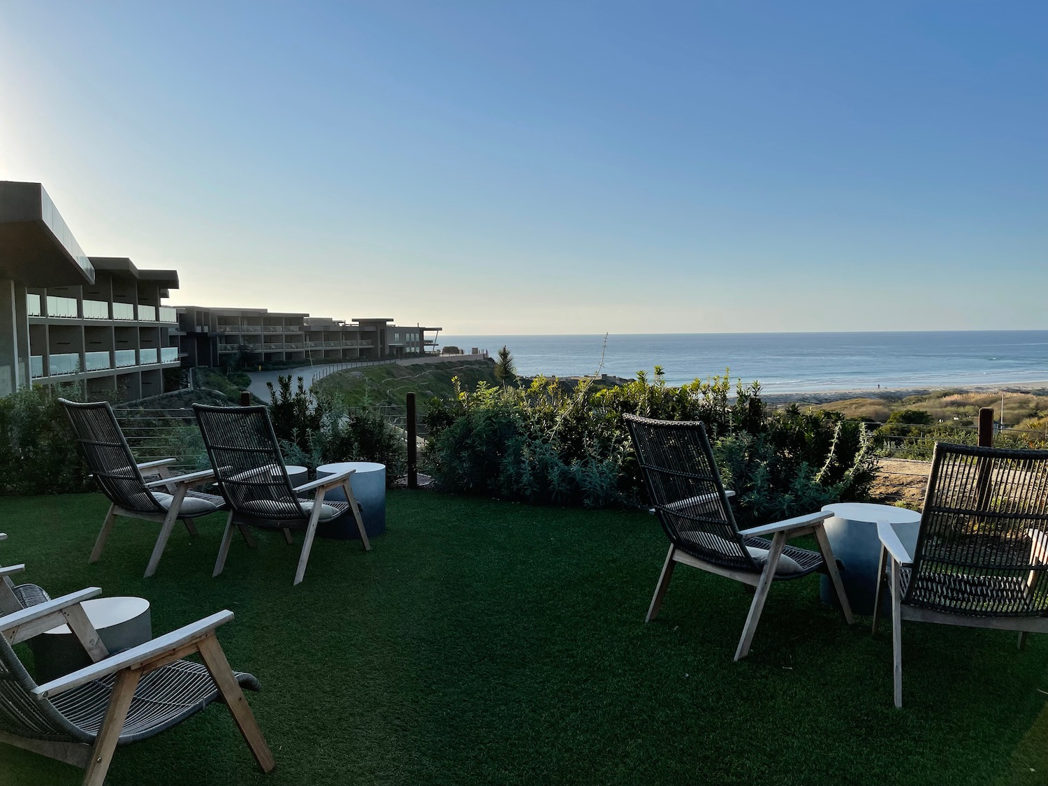 a patio with chairs and tables on a grassy area with a beach and ocean