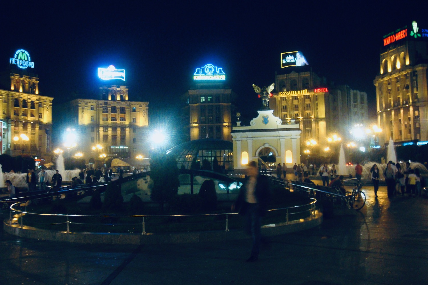 a group of people walking in a plaza with buildings and a fountain