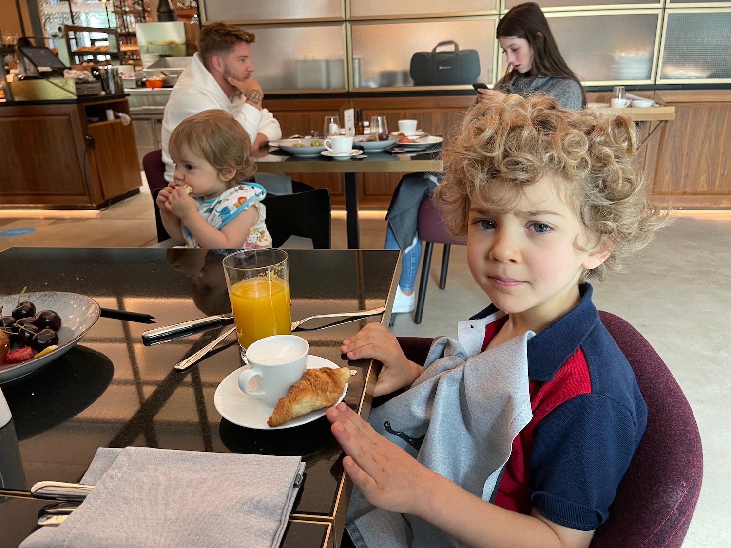 a boy sitting at a table with a croissant and a glass of juice