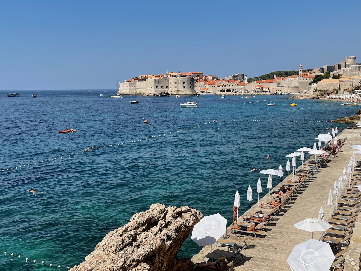 a beach with umbrellas and a body of water with a stone walkway and buildings in the background