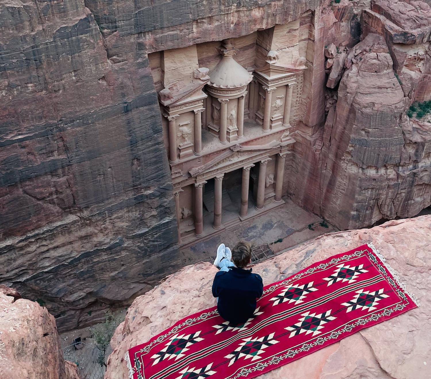 a person sitting on a red rug looking at a building in a canyon