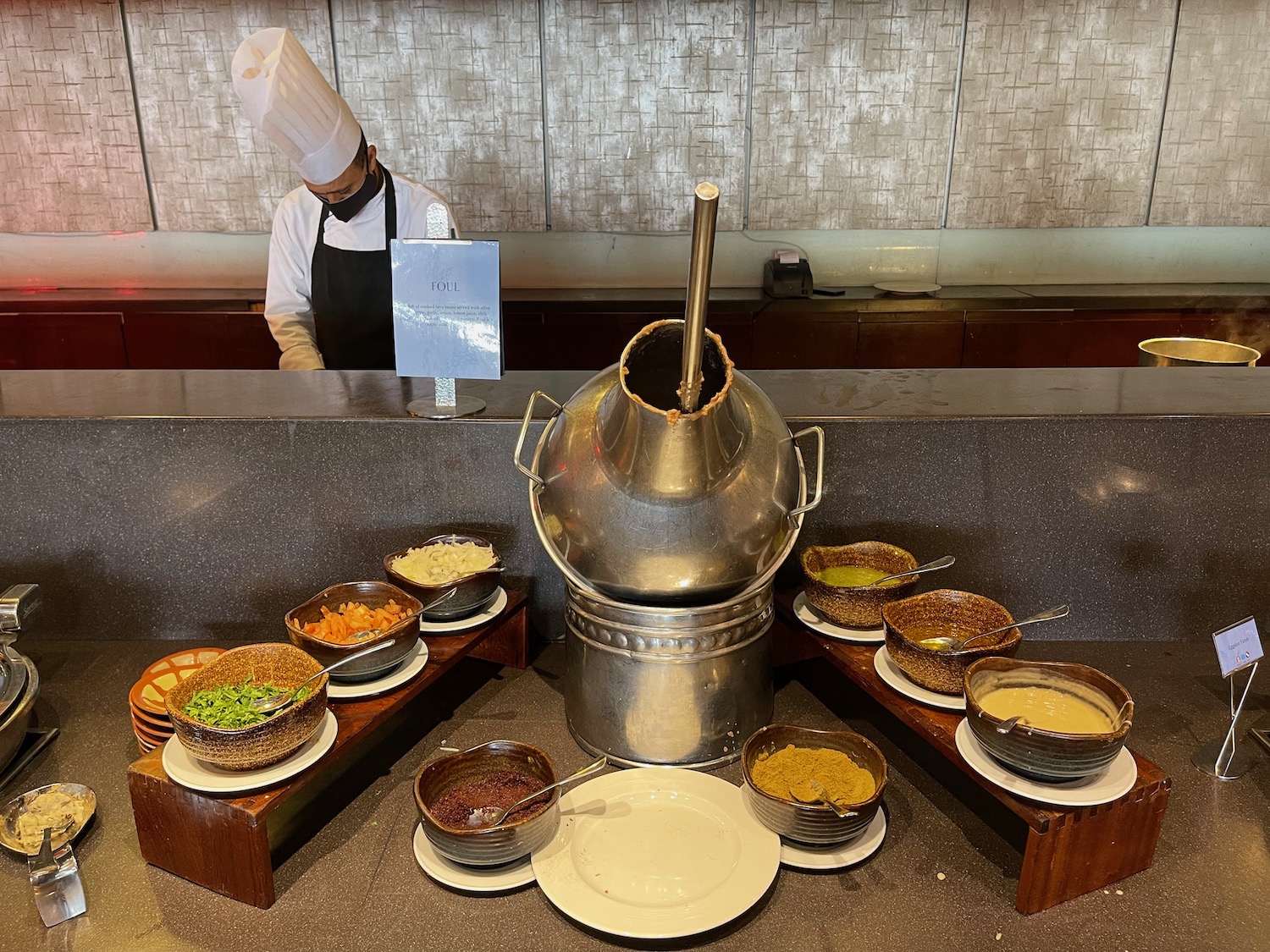 a chef standing behind a counter with a silver pot and bowls of food