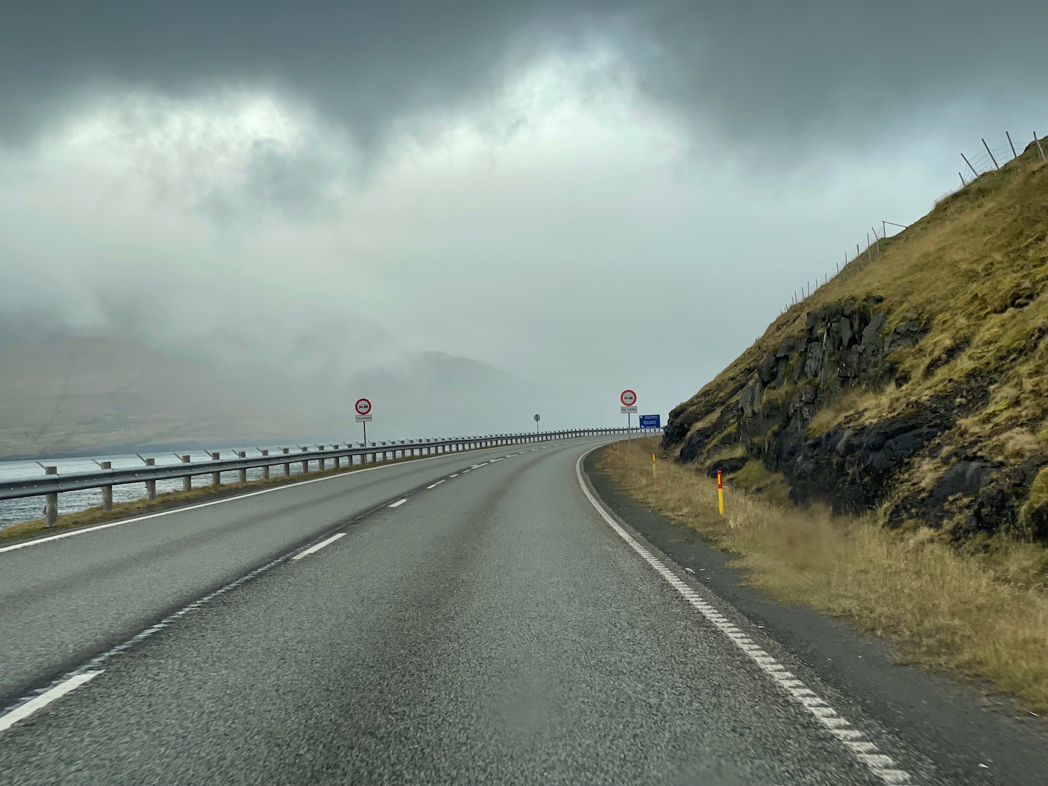 a road with a railing and a hill with a body of water and clouds