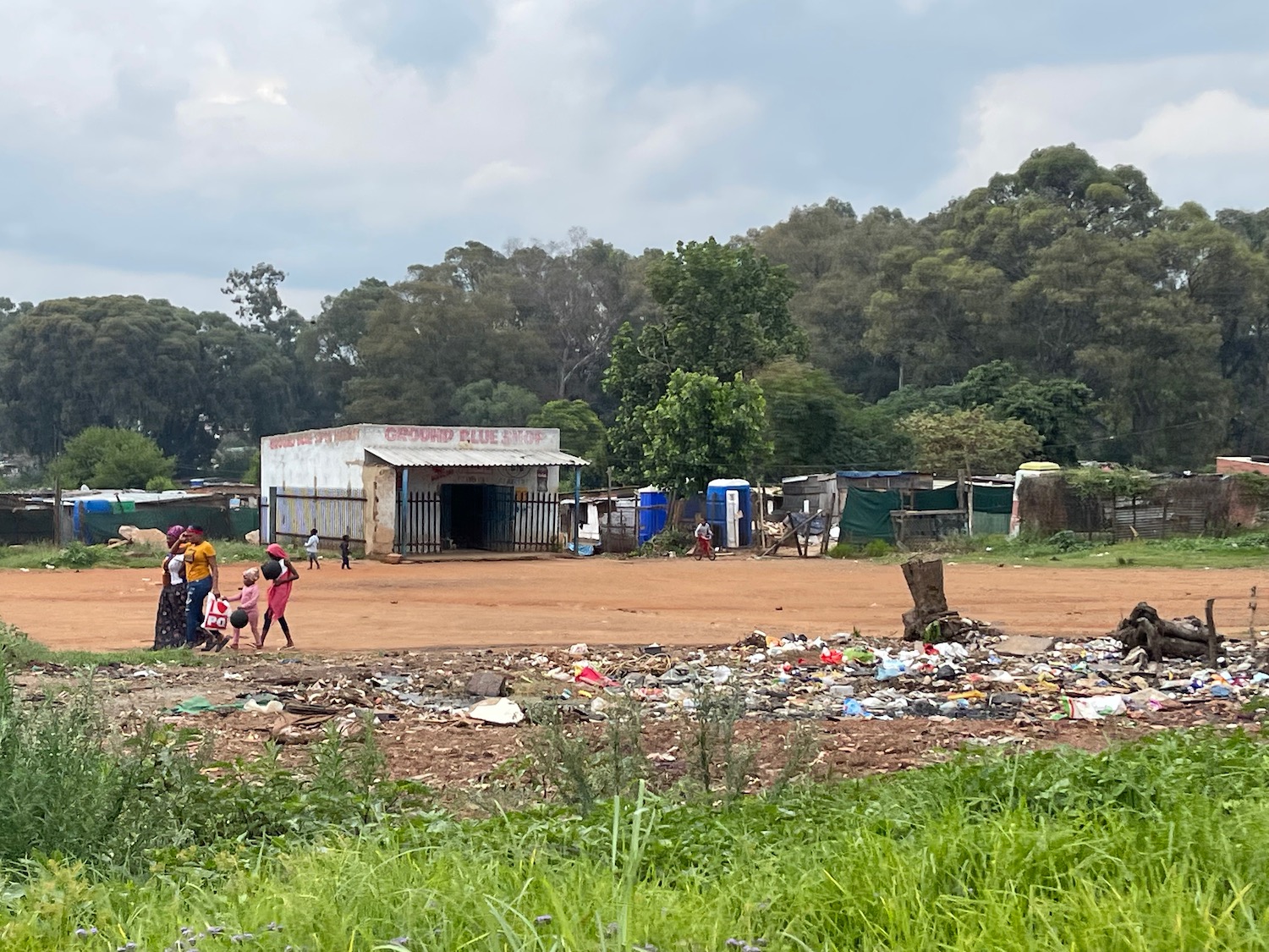 a group of people standing in a dirt field with trash and a building