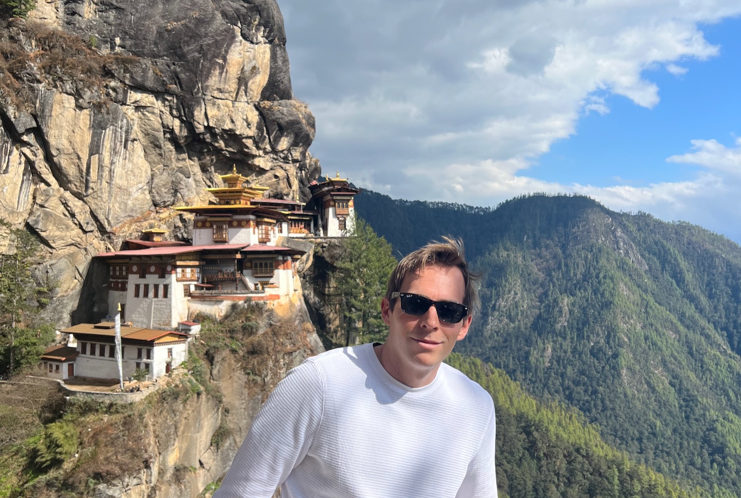 a man standing on a cliff with buildings on the side with Paro Taktsang in the background