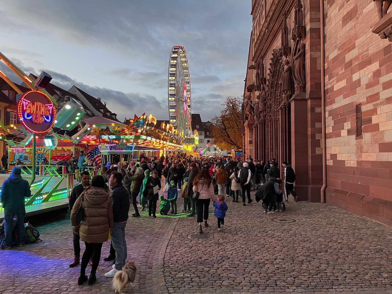 a crowd of people walking on a cobblestone street with a ferris wheel