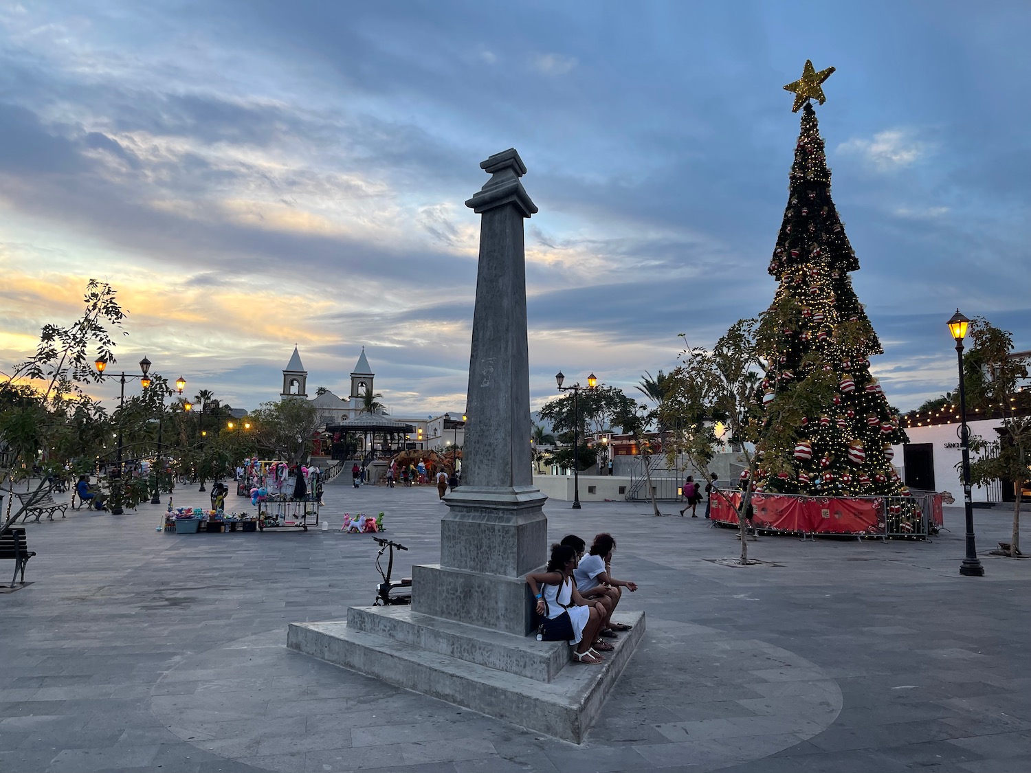 a group of people sitting on a stone pillar in a plaza with a christmas tree
