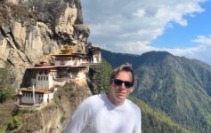 a man standing on a cliff with buildings on the side with Paro Taktsang in the background