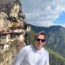 a man standing on a cliff with buildings on the side with Paro Taktsang in the background