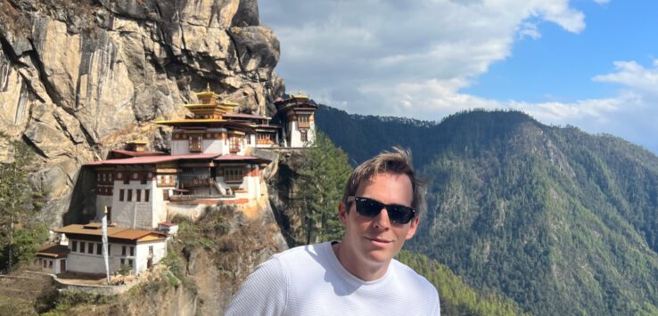 a man standing on a cliff with buildings on the side with Paro Taktsang in the background