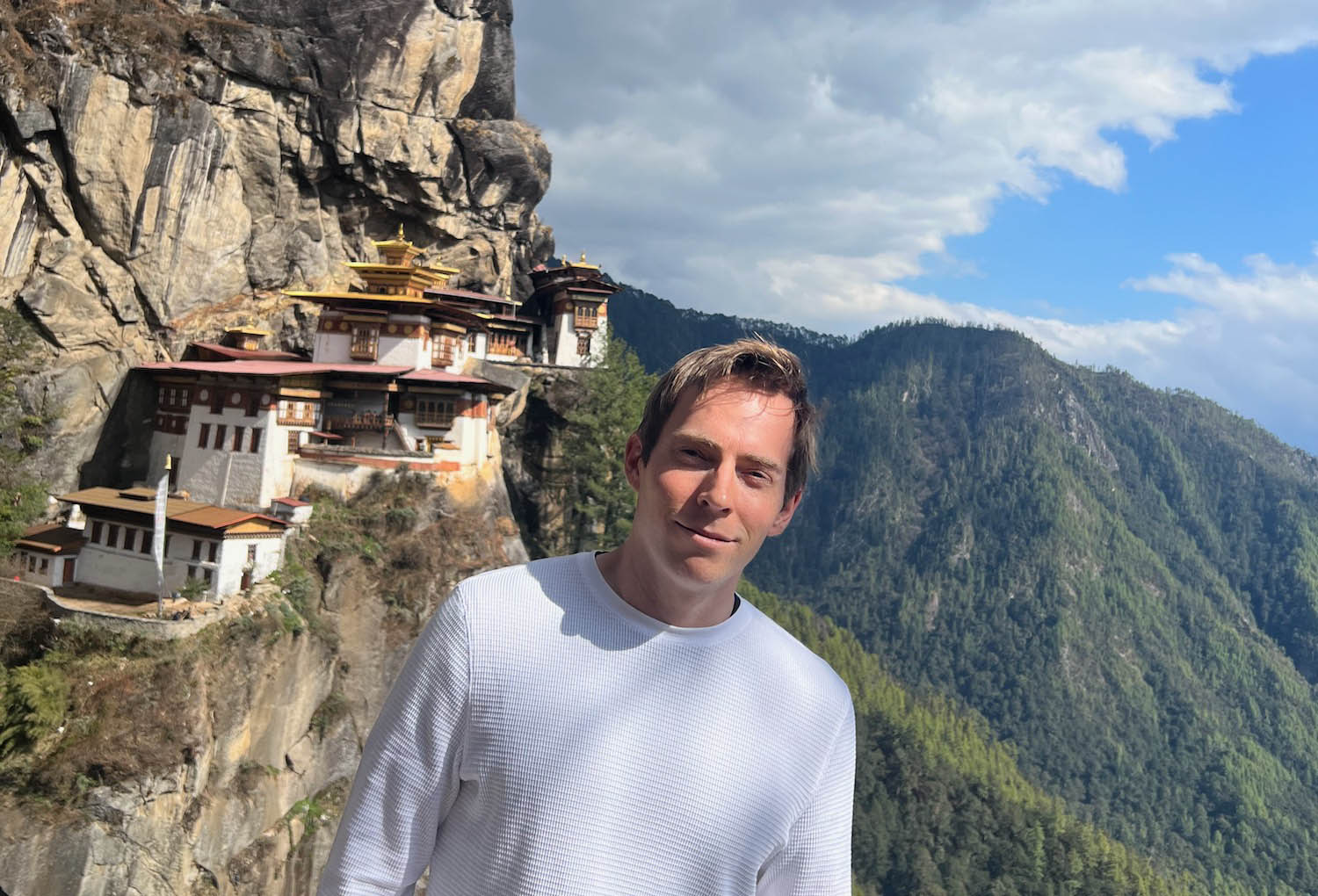 a man standing on a cliff with buildings on the side with Paro Taktsang in the background