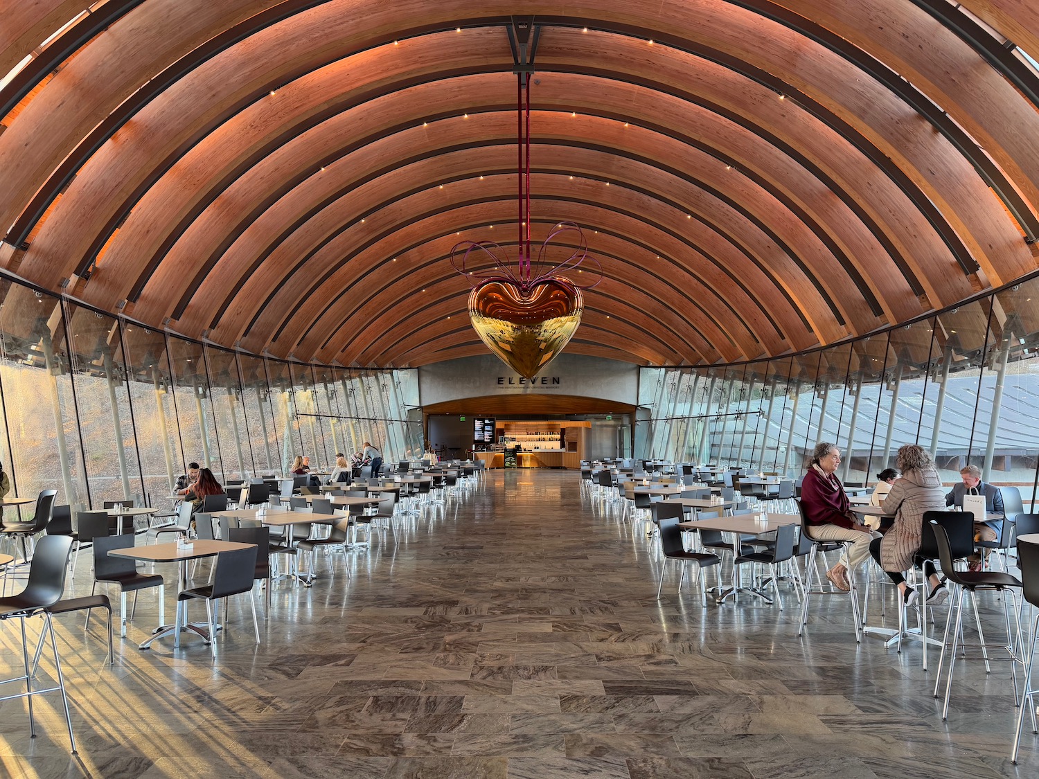 a room with tables and chairs and a large gold heart from the ceiling with Crystal Bridges Museum of American Art in the background