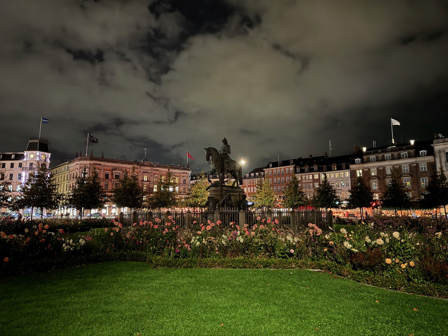 a statue in a park with flowers and buildings in the background