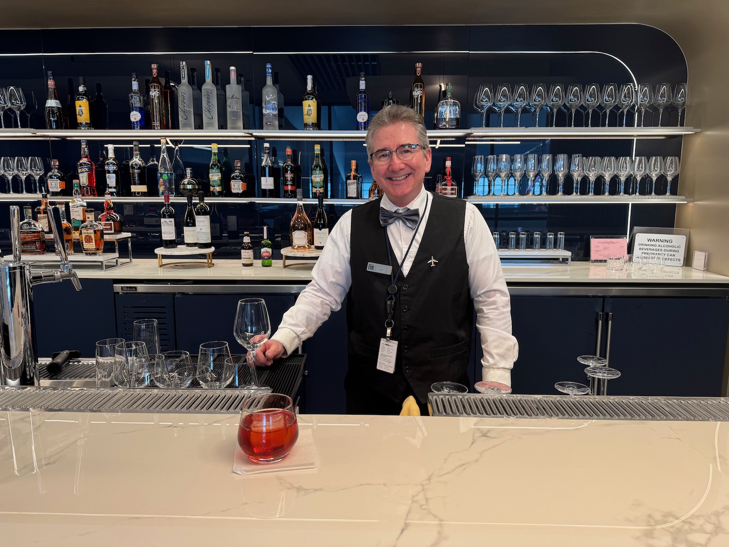 a man standing behind a counter with glasses and a bar full of wine