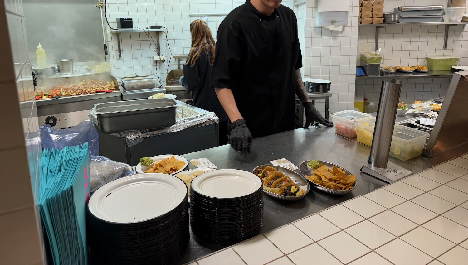 a man in a black shirt and gloves standing in a kitchen with plates and food