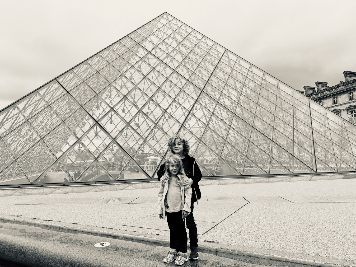 two children standing in front of a glass pyramid with Louvre in the background