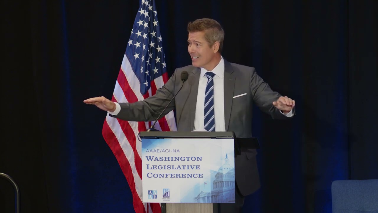 a man standing at a podium with a microphone and a flag behind him