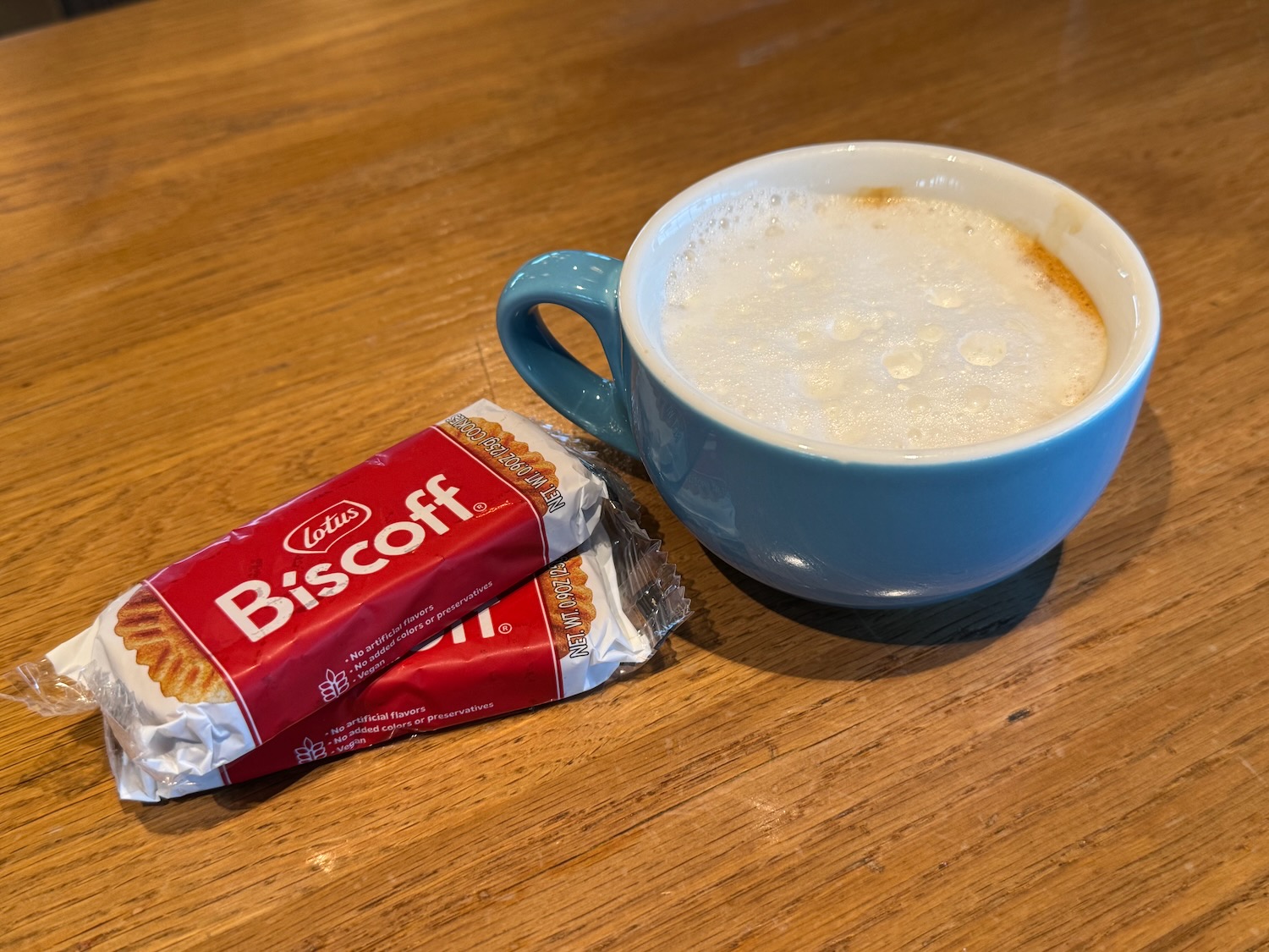 a blue mug with foam and a red packet of biscoff on a wood table